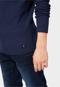 Pull texturé bleu marine à manches longues, avec des poignets côtelés et une petite étiquette de logo sur l'ourlet. Porté avec un jean en denim bleu foncé.