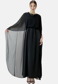 Robe longue noire avec manches cape transparentes, tissu fluide, taille froncée et silhouette élégante. Associée à des sandales noires à brides.