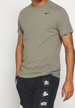 Homme portant un t-shirt Nike vert olive et un short noir avec des imprimés graphiques blancs sur le côté gauche, debout avec une main dans la poche.
