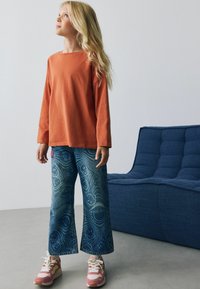 Tricou din bumbac cu mânecă lungă, de culoare portocalie ruginită, asortat cu pantaloni largi albaștri cu un model spiralat, completat cu adidași multicolori.