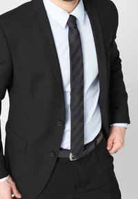 Czarny garnitur o dopasowanym kroju, biała koszula do garnituru i czarny krawat w paski. Posiada srebrną klamrę oraz fakturowany materiał.