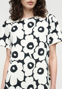 Γυναίκα που φοράει ασπρόμαυρο φόρεμα με κοντά μανίκια και μεγάλο αφηρημένο σχέδιο λουλουδιών, εμφανιζόμενη από τους ώμους μέχρι τη μέση.