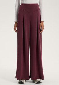 Pantaloni largi violet, cu talie înaltă, care dispun de pliseuri verticale și o textură netedă și moale. Purtați cu adidași în nuanțe neutre.