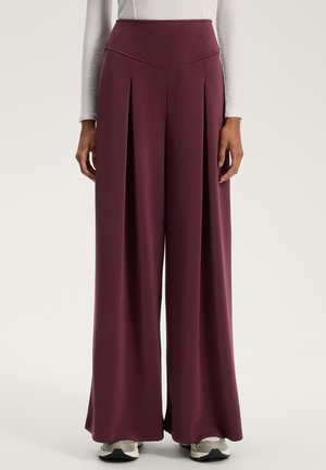 Pantaloni largi violet, cu talie înaltă, care dispun de pliseuri verticale și o textură netedă și moale. Purtați cu adidași în nuanțe neutre.