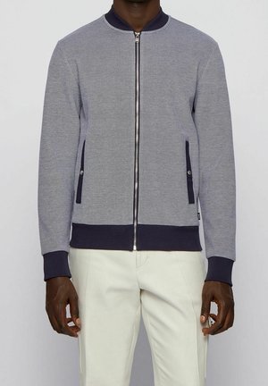 Mann trägt eine hellgraue Jacke mit Reißverschluss sowie dunkelblauen Bündchen, Kragen, Saum und Taschenabschlüssen, kombiniert mit weißen Hosen.