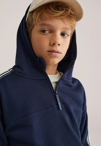 Tumši zilas krāsas kapučjakas ar puspašjostu dizainu, kurai ir teksturēta auduma virsma, priekšējā kabata un baltas svītras uz piedurknēm.