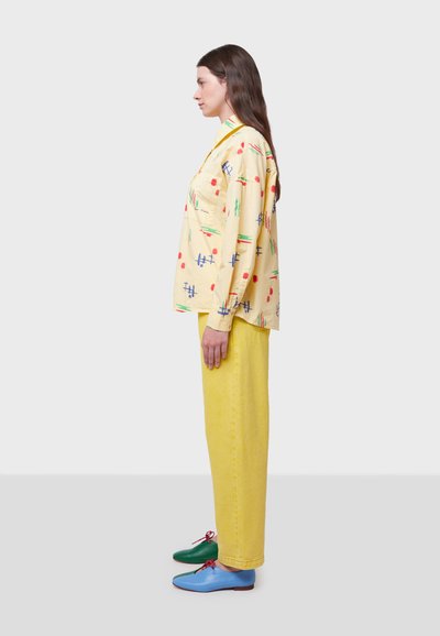 Camisa amarilla con estampado de diseños geométricos rojos y azules, combinada con pantalones de mezclilla amarillos de cintura alta; lleva zapatos verdes y azules.