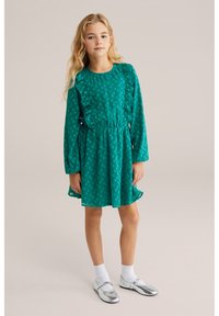 Zaļa kleita ar teksturētu punktiņu rakstu, volāniem pie izgriezuma un piedurknēm, elastīgu vidukli, kombinēta ar sudraba apaviem un baltām zeķēm.