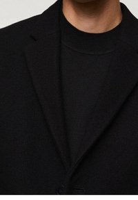 Blazer en mélange de laine noire avec revers crantés, fermeture à un bouton, texture lisse, et un col roulé côtelé noir en dessous.