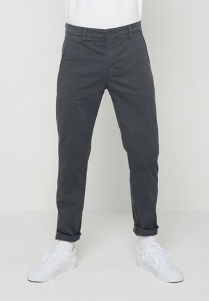 Man draagt donkergrijze slim-fit broek met omgeslagen pijpen bij de enkels en witte sneakers tegen een effen lichte achtergrond.