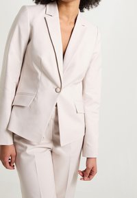 Vaaleanpunainen bleiseri, jossa yksi nappikiinnitys, loviolkaimet ja kaksi etutaskua. Sileä rakenne ja räätälöity istuvuus.