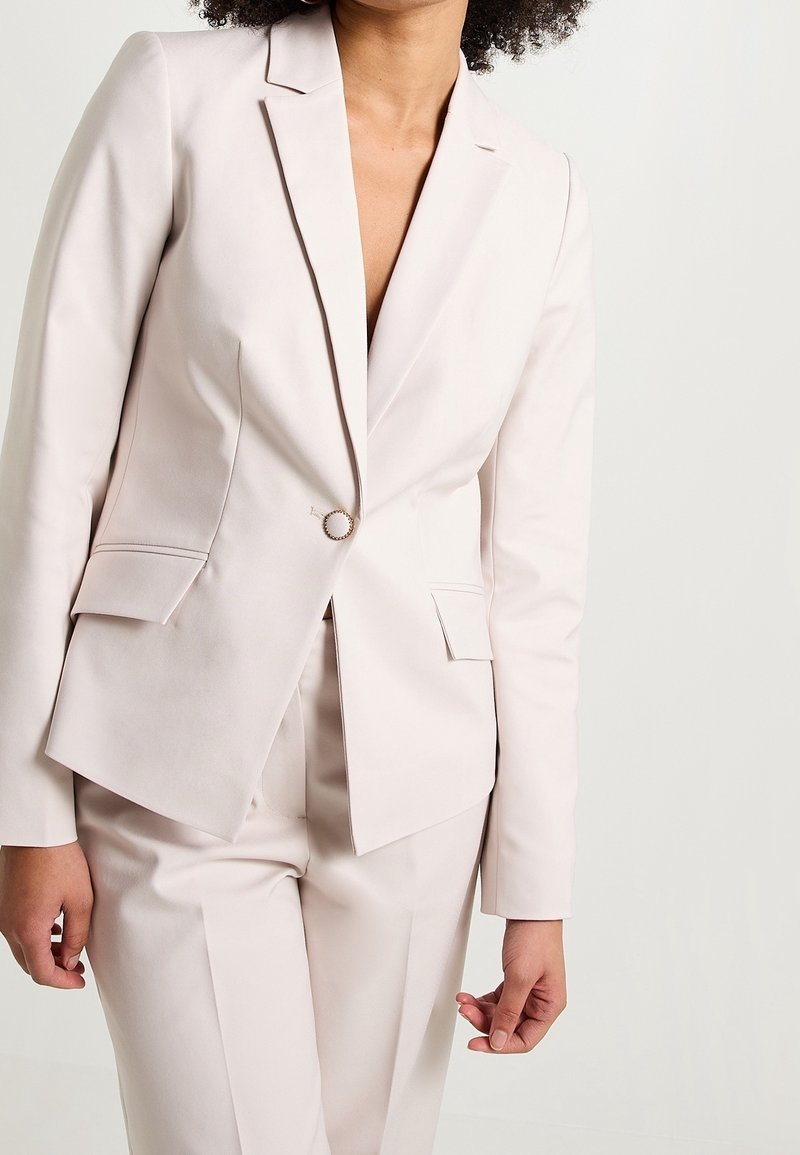 Vaaleanpunainen bleiseri, jossa yksi nappikiinnitys, loviolkaimet ja kaksi etutaskua. Sileä rakenne ja räätälöity istuvuus.