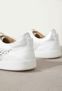 Weiße Sneakers mit einer strukturierten Lederoberfläche, silbernen Akzenten an der Ferse und einer geflochtenen Detailverzierung an den Seiten. Gummisohle sichtbar darunter.
