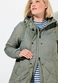 Blonde vrouw in een groene gewatteerde jas over een blauw en zwart gestreept shirt, die de rits van de jas aanpast.
