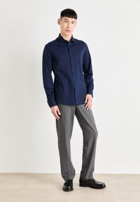 Jūras flotes ilga piedurkne krekls ar pogu slēgšanu, savienots ar gaiši pelēkiem biksēm un melniem uzvalka apaviem, uz neitrālas fona.