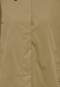 Trench-coat beige avec fermeture boutonnée dissimulée et deux poches latérales, montrant les détails de la partie avant au niveau du torse.