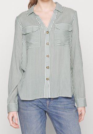 Person iført en grøn og hvid lodret stribet skjorte med to forlommer, kombineret med blå jeans.