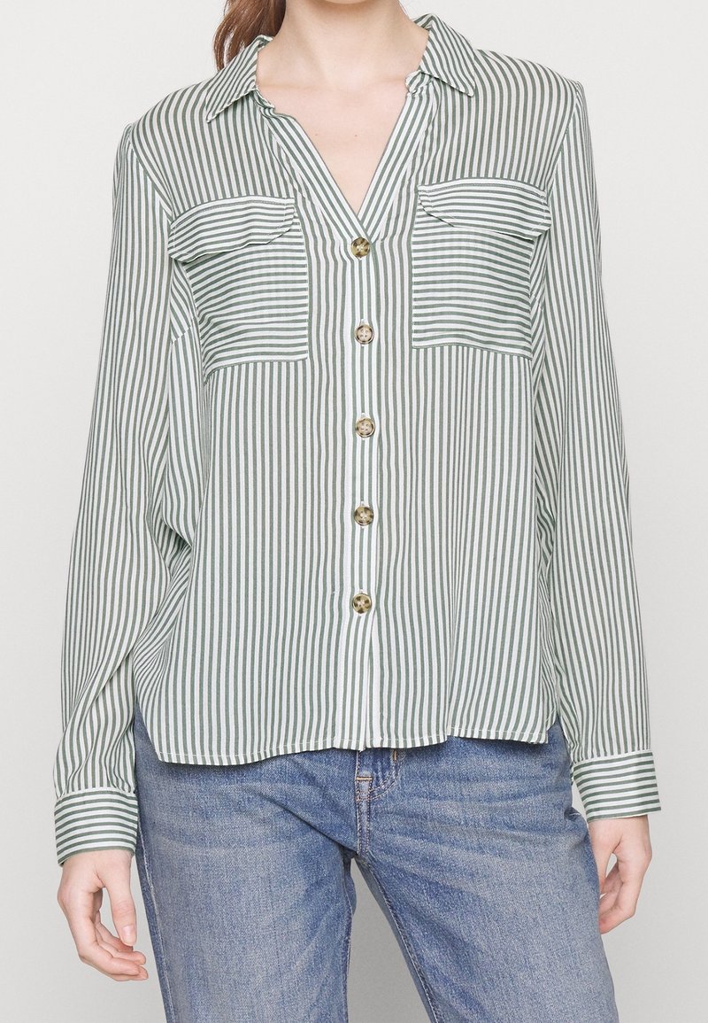 Personne portant une chemise boutonnée à rayures verticales vertes et blanches avec deux poches avant, associée à un jean bleu.