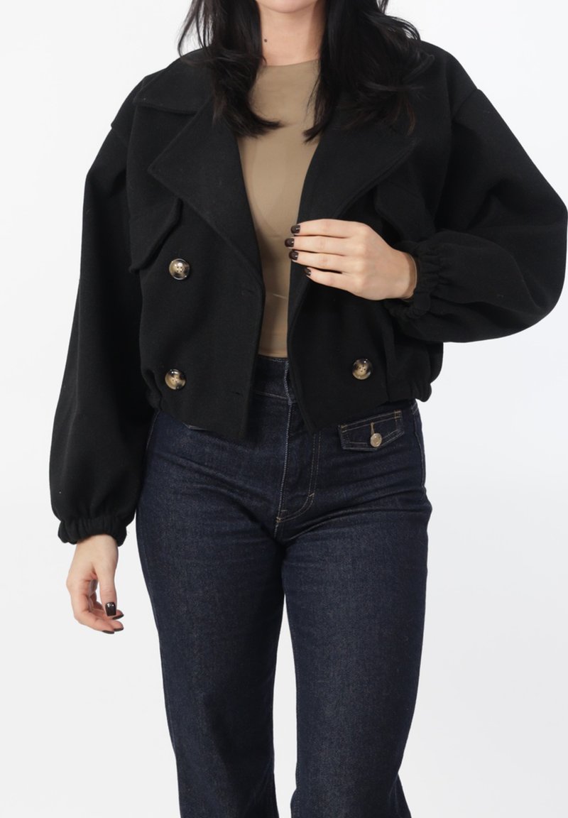 Zwarte cropped jas met grote knopen, brede revers en geplooide mouwen. Draagt over een beige top en gecombineerd met donkeren denim jeans.