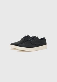 Zapatillas de ante negro con una suela de goma color crema. Las características incluyen una punta redonda, un diseño de cordones sencillo y acentos de costura mínimos.