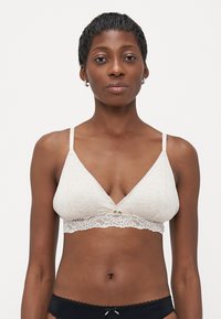Lichtbeige bralette met een kanten rand aan de onderkant, heeft een driehoekige vorm, verstelbare bandjes en een klein decoratief knoopdetail.