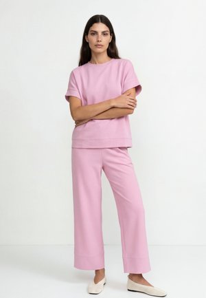 Nő áll összefont karokkal, világos rózsaszín rövid ujjú felsőt és bőszárú nadrágot visel, fehér slip-on cipővel.