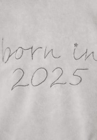 Ljusgrått tyg med broderad text som säger "född 2025" i en enkel, kursiv stil med en stjärnaccent, mjuk textur.