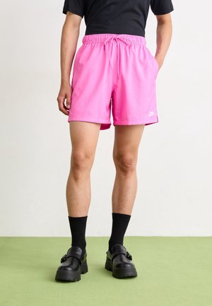 Personne portant un short Nike rose vif avec cordon, un t-shirt noir, des chaussettes noires et des mocassins noirs épais, debout sur un sol vert.