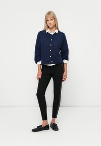 Navy blue cardigan with buttons and puffy sleeves, paired with black slim-fit pants featuring a white side stripe and flat black shoes.