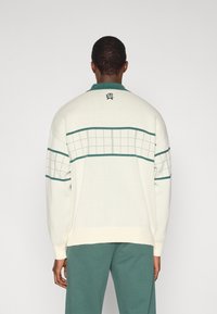 Pull en maille blanche avec des accents verts, présentant un motif en grille sur le dos et un col à revers. Texture douce et coupe décontractée.