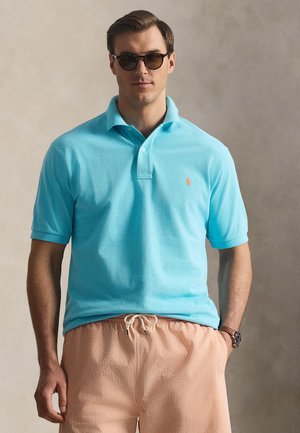 Mees, kellel on seljas türkiissinine polomehe särk, oranžid paelaga lühikesed püksid, päikeseprillid ja käekell, seisab neutraalse tausta ees.