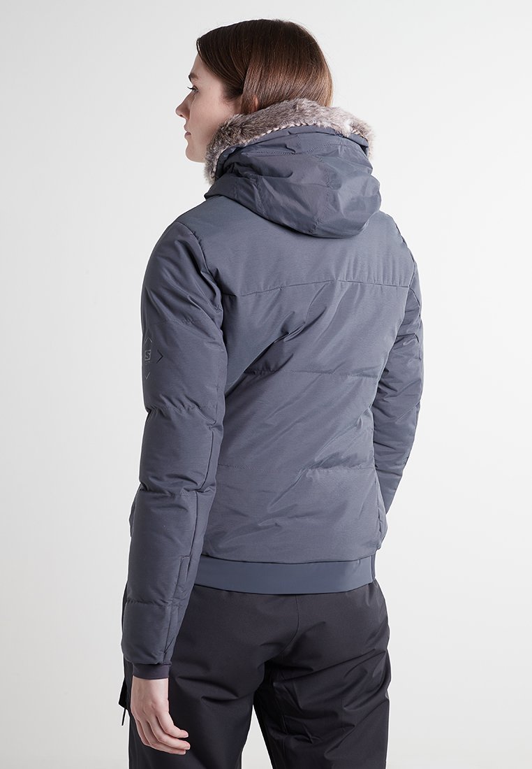 Chaqueta acolchada gris con capucha forrada de piel, diseño acolchado y puños elásticos. Presenta un exterior suave y una silueta ajustada.