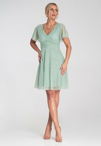 Mintgrünes Kleid mit kurzen, durchsichtigen Ärmeln und einem taillierten Oberteil. Ausgestellter Rock mit schichtiger Textur, kombiniert mit beigen, hohen Sandalen mit Riemchen.