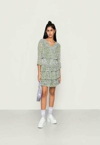 Mulher jovem em vestido curto verde com padrão e sapatos de plataforma brancos, segurando uma pequena bolsa de lavanda, de pé contra um fundo simples.