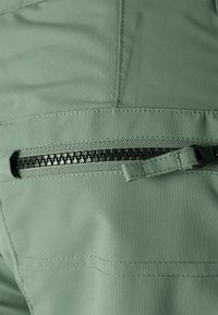 Zelená látka s hladkým povrchem, na které jsou černý zip a švy. Zip je částečně rozepnutý, což naznačuje kapsu.