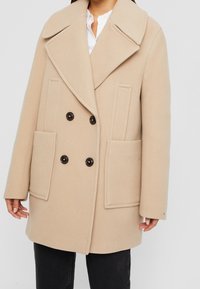 Abrigo beige de doble botonadura con grandes solapas, que cuenta con dos bolsillos frontales y botones oscuros, hecho de una tela suave y texturizada.