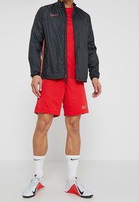 Chaqueta cortavientos Nike en negro con acentos rojos, combinada con una camiseta roja de manga corta y pantalones cortos rojos. Calcetines blancos y zapatillas gris/rojas completan el look.