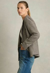 Blazer s potpisom čavrljanja u smešnoj smeši smešnog i krema, teksturirana tkanina, otvoreni prednji deo, srednje dužine rukava, uparen s plavim farmerkama. Neutralna pozadina.