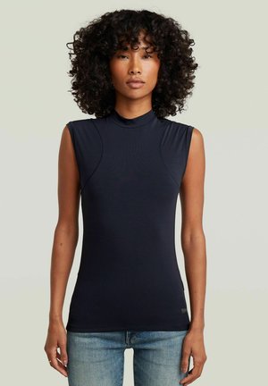 Donna con capelli ricci che indossa un top nero aderente senza maniche e jeans blu, in piedi davanti a uno sfondo chiaro e neutro.