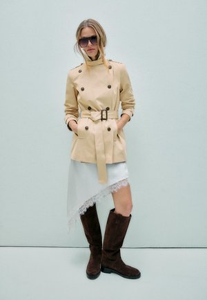 Ženska, oblečena v bež trenč plašč z pasom, belo asimetrično čipkasto krilo, temno rjava škornji do kolen in velika sončna očala, stoji ob svetlem zidu.