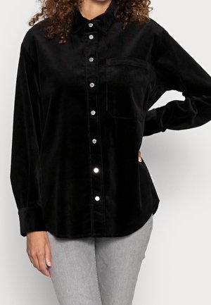Camicia di velluto a coste nera con chiusura a bottoni, maniche lunghe, colletto e una tasca sul petto. Presenta bottoni a pressione argentati e orlo curvo.