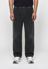 Μαύροι παντελόνια από κορντόριο με άνετη εφαρμογή, φορεμένα με λευκά και γκρι αθλητικά παπούτσια, σε όρθιο άτομο μπροστά σε απλό φόντο.