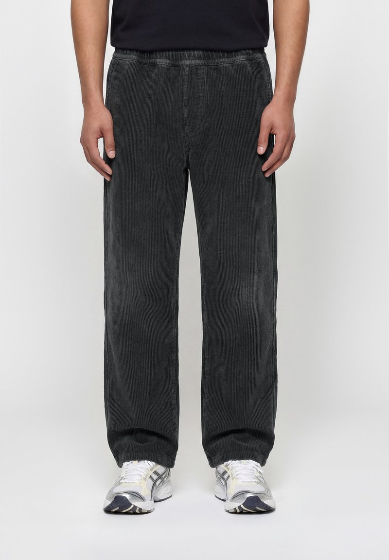 Μαύροι παντελόνια από κορντόριο με άνετη εφαρμογή, φορεμένα με λευκά και γκρι αθλητικά παπούτσια, σε όρθιο άτομο μπροστά σε απλό φόντο.