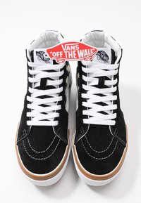Zapatillas altas de ante negro con cordones blancos y suela de goma. Presenta la icónica etiqueta "Vans Off the Wall" en la parte superior. Parte superior texturizada y acentos cosidos.