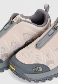 Двойка бежови и сиви туристически обувки The North Face с предно закопчаване с цип и подметки Vibram, показващи детайли отблизо и текстурирана материя.