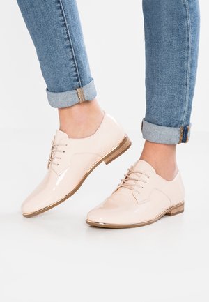 Stopy w błyszczących beżowych oxfordach z sznurowadłami w zestawieniu z podwiniętymi niebieskimi dżinsami na białej podłodze.