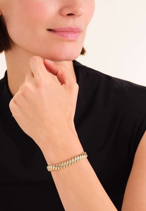 Pulsera de oro con un diseño retorcido, con un acabado metálico suave. Lleva puesta en una muñeca, mostrando su superficie lisa y reflectante.