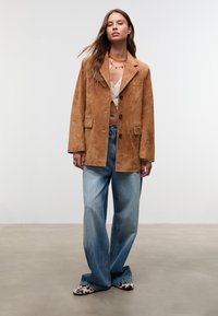 Kvinde iført en brun ruskindsblazer, en hvid, kortærmet knap-top, blå vide jeans, sko med kvægprint og lagdelte perlekæder, der står på et beton gulv.