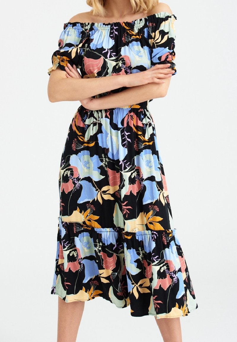 Femme portant une robe midi épaules dénudées avec taille élastique et grand imprimé floral coloré sur fond noir.