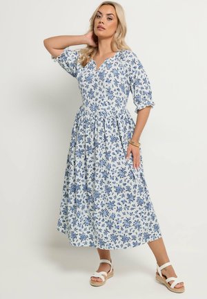 Kvinde iført en blå og hvid midikjole med floralmønster, pufærmer og bølget udskæring, poserer med den ene hånd bag hovedet og sandaler.